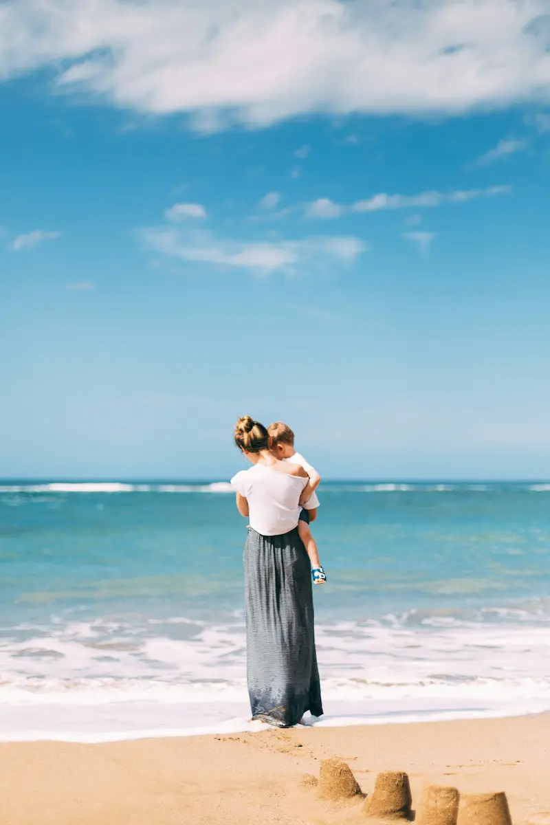 A woman holds a child on a beach. Photo from the back.