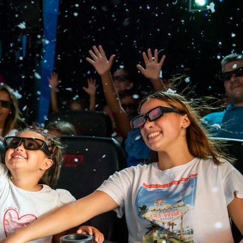 孩子们和成年人戴着3D眼镜坐在剧院中，微笑着举起双手，周围飘落着彩纸或雪花。