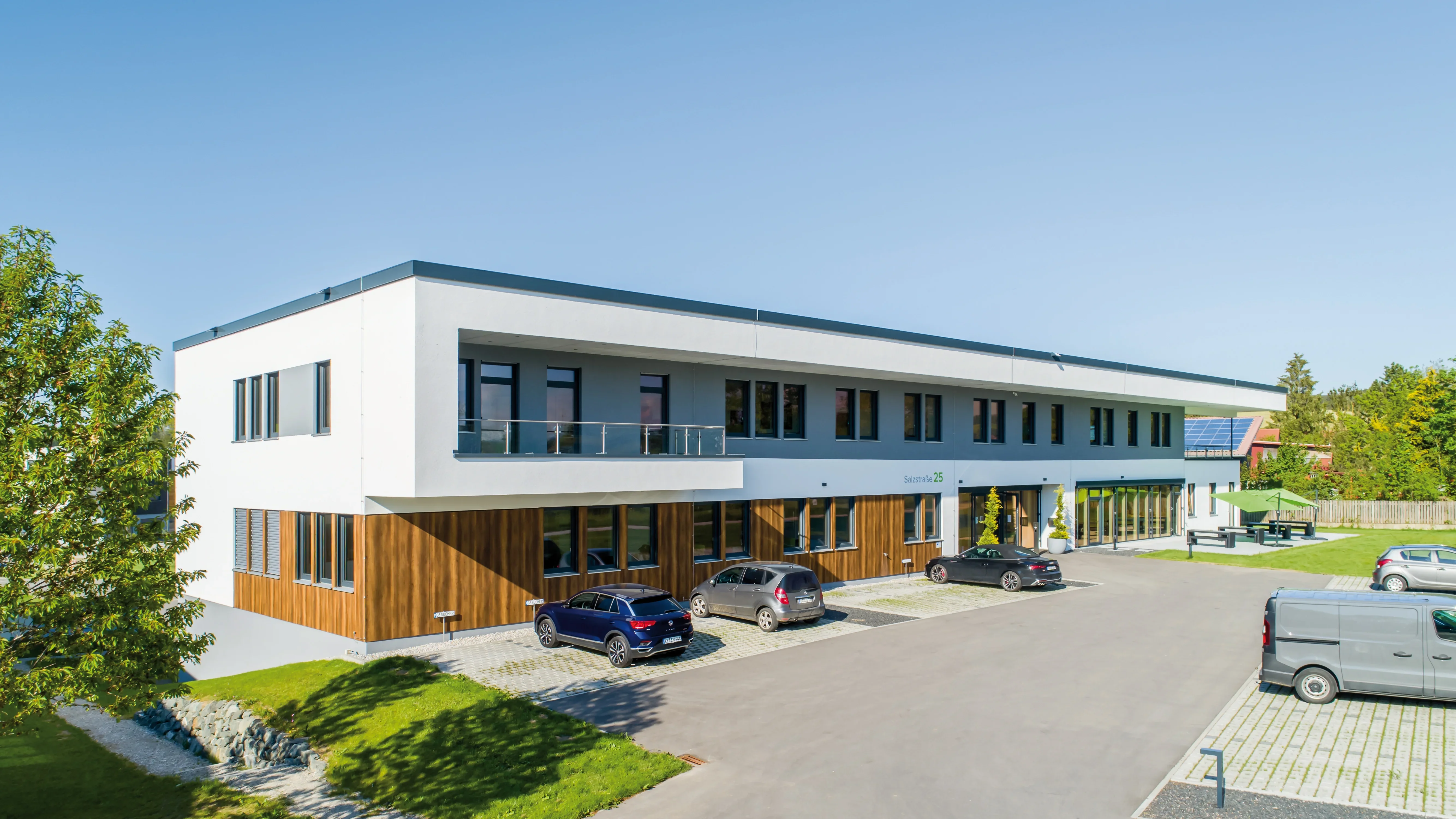 Modernes Bürogebäude mit Holzverkleidung, grauem Dach und Balkon. Mehrere Fahrzeuge parken vor dem Gebäude an einem sonnigen Tag.