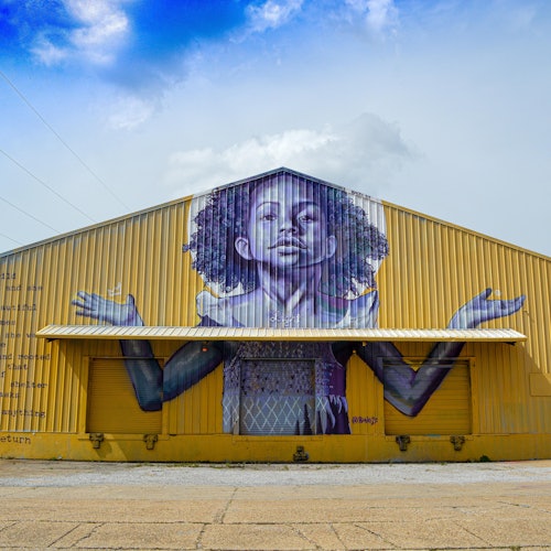 在部分多云的天空下，一个黄色仓库上画着一个大幅壁画：一个长着卷发的小孩，双臂伸展开来。