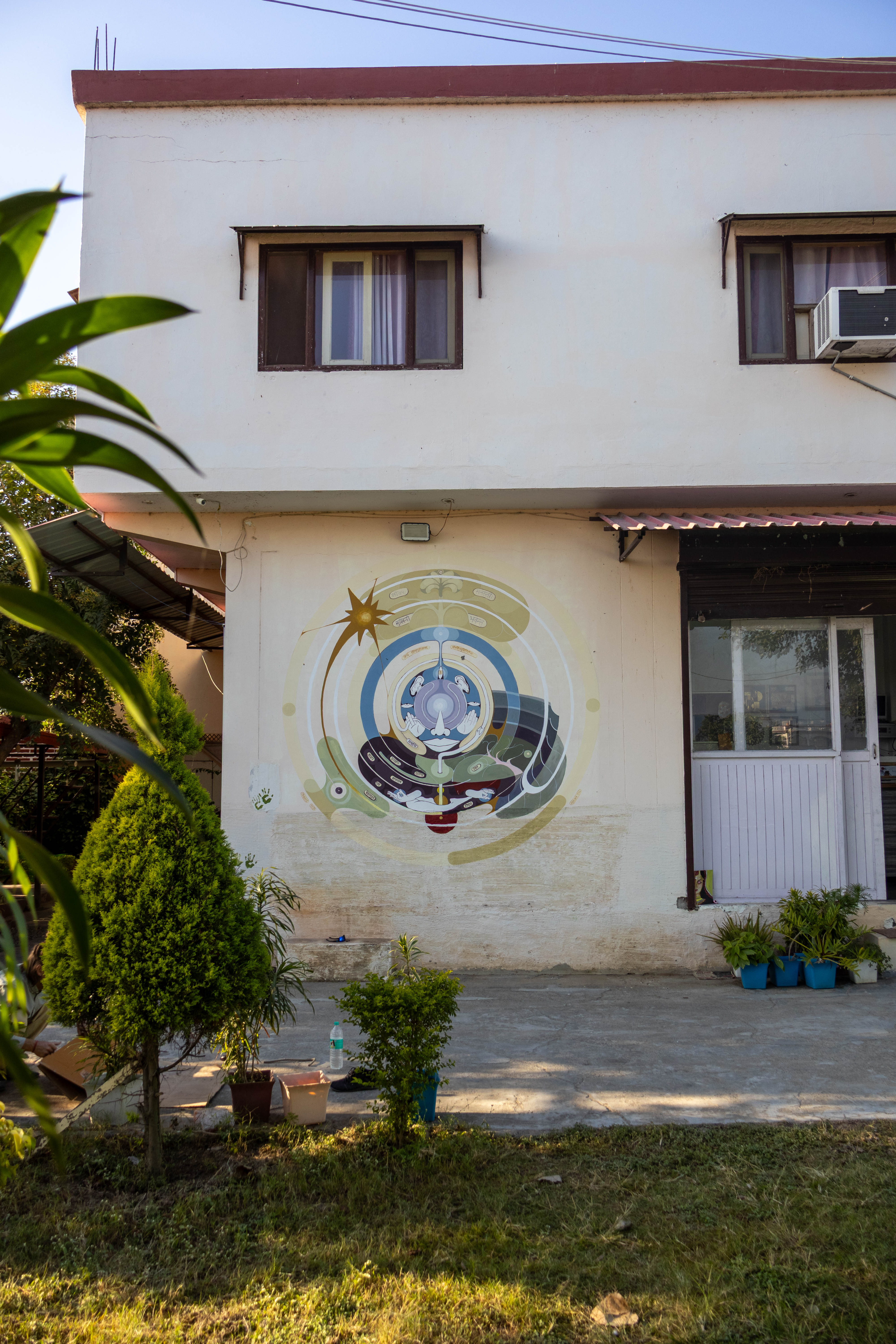 A circular mandala-style mural on a white building near the entrance surrounded by several potted plants next to a garden.