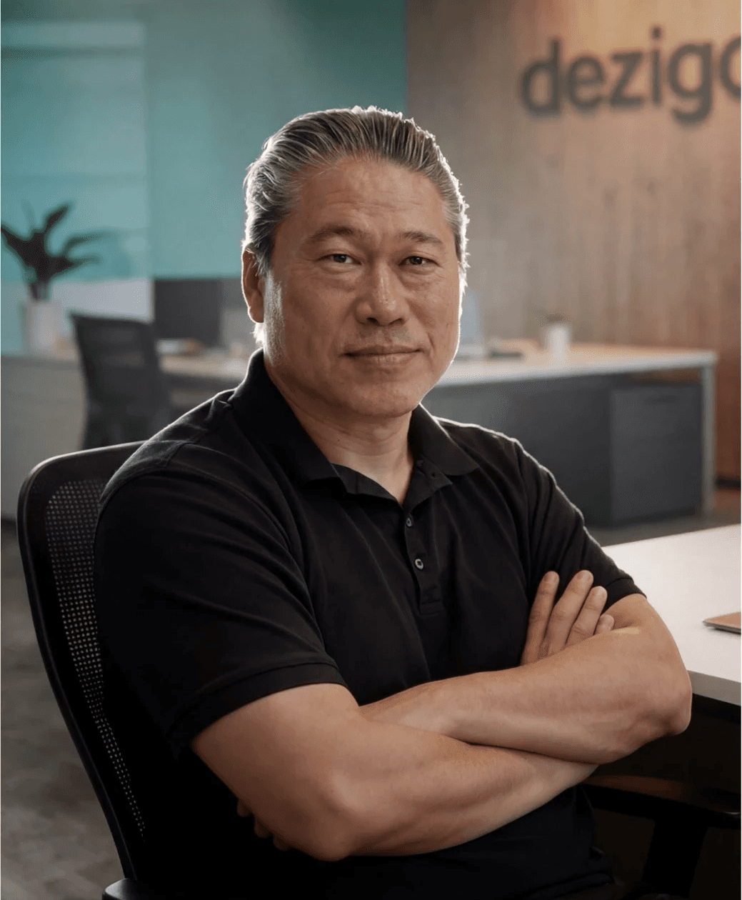 A person with blond hair wears a dark coat and a beige turtleneck sweater. They have a calm expression, against a neutral background.