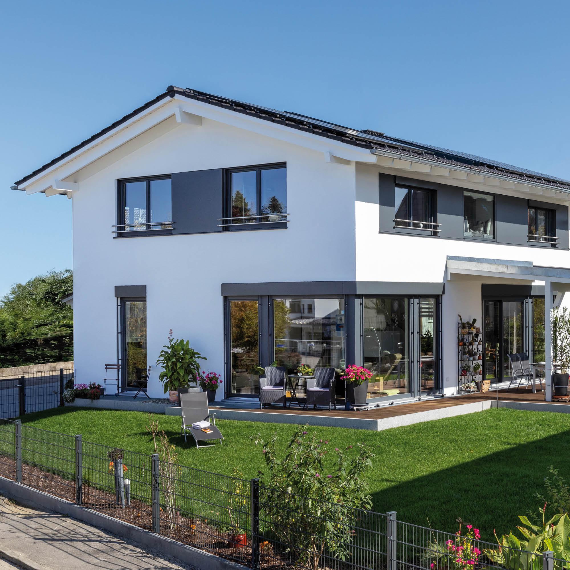 Modernes weißes Einfamilienhaus mit dunklen Akzenten, Solaranlage auf dem Dach und großer Terrasse mit Sitzmöbeln.