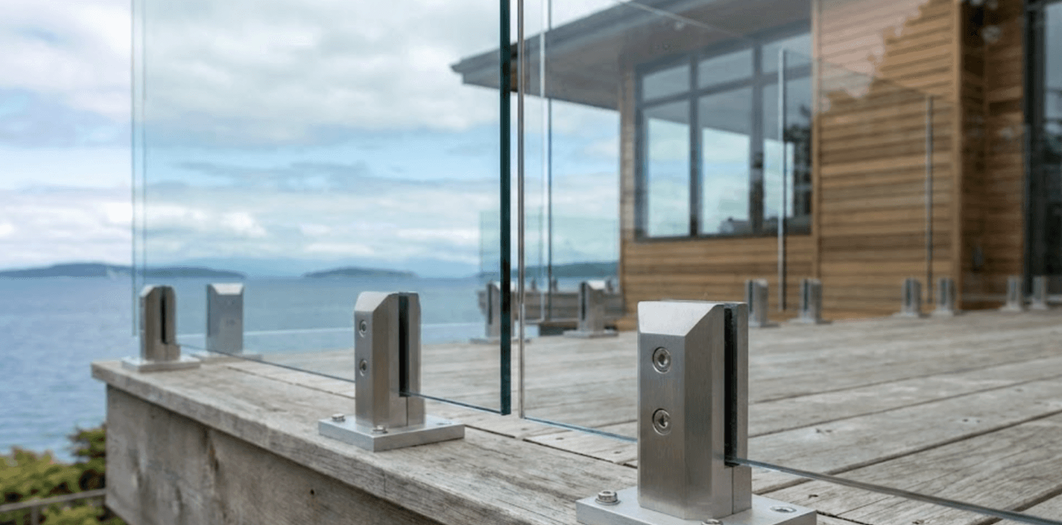 Close-up detail shot of frameless glass railing installation on White Rock waterfront deck, showing aluminum base clamp mounting hardware, ocean view visible through glass panels, professional craftsmanship, modern coastal home exterior"