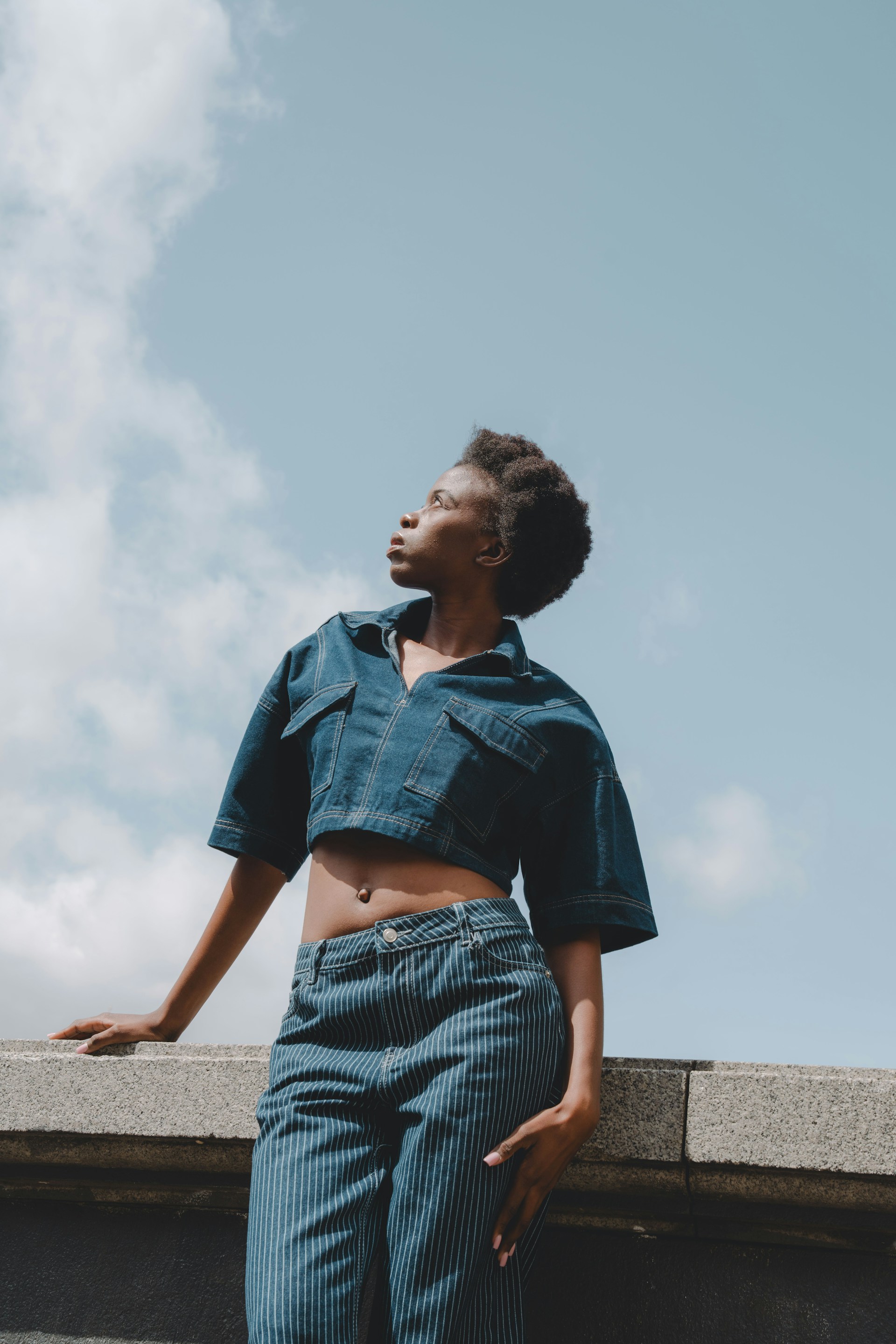 The woman posing against a stone ledge, captured from a low angle against a vast blue sky. She is looking to the side, highlighting the pinstripe detail of her trousers and the cropped fit of her denim top.