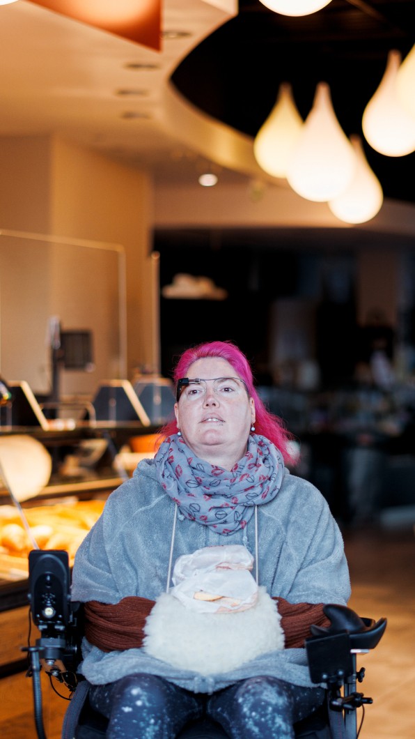 Saskia, a user of the munevo smart glasses, sitting in a motorized wheelchair inside a café. She has striking pink hair and is wearing a grey scarf and a light grey jacket. Saskia is looking forward, using the smart glasses to navigate or interact with her environment. The café setting is warmly lit, with stylish hanging lights in the background, creating a cozy and inviting atmosphere. This setting emphasizes the everyday usability of the munevo glasses, enabling mobility and independence for individuals in various social settings