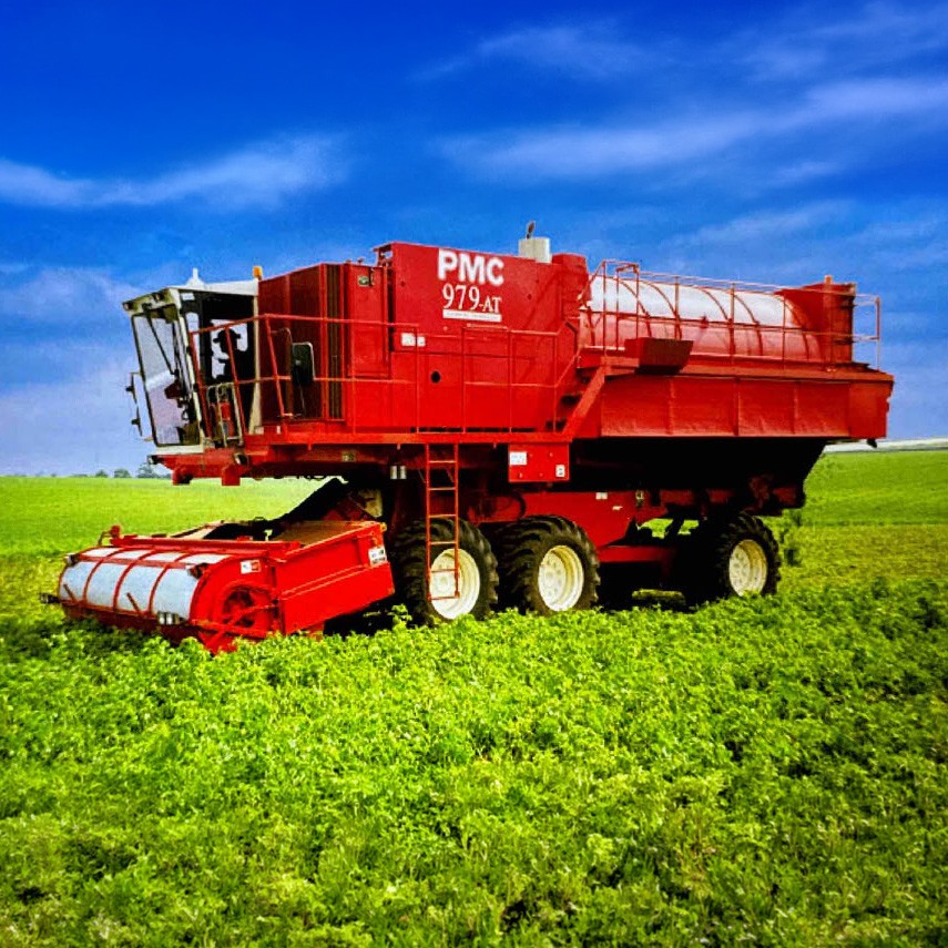 A red PMC 979-AT self-propelled pea harvester working through a lush green pea crop under a bright blue sky.