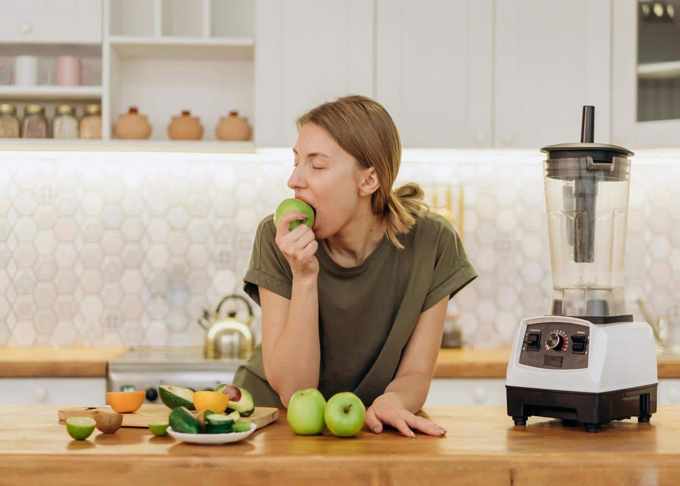 Woman eating green apple with veggies and a blender on the counter in front of her