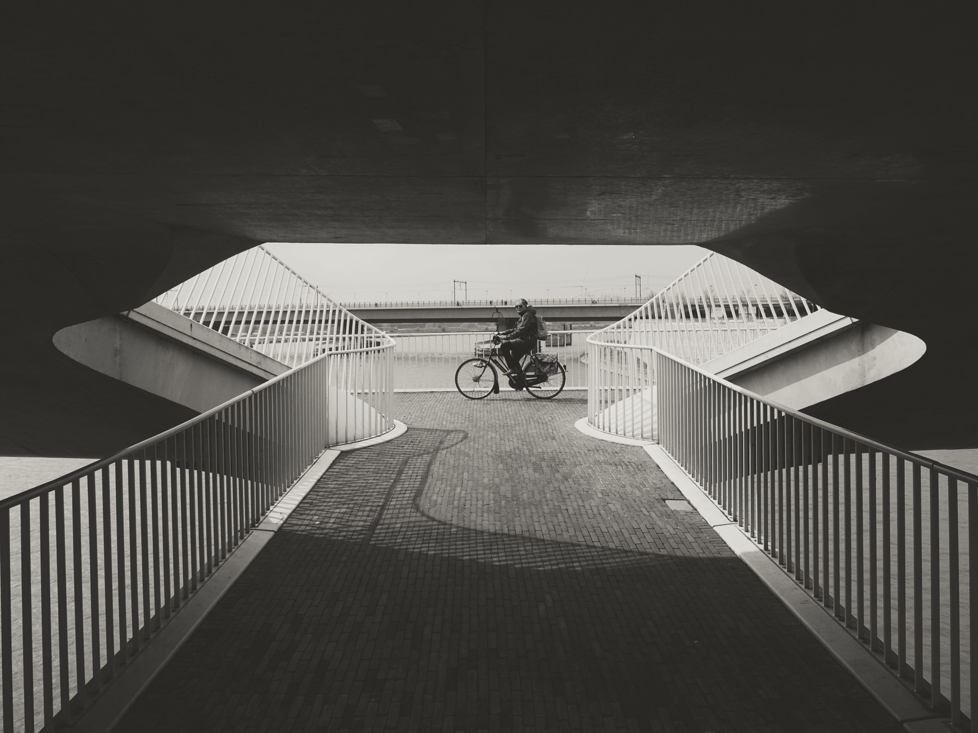 Cyclist on bike path under bridge with metal railings and brick pavement in urban setting.