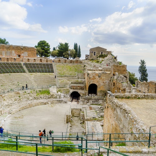 Ancien amphithéâtre en pierre avec peu de visiteurs, entouré de ruines, de verdure, et surplombant une mer lointaine sous un ciel partiellement nuageux.