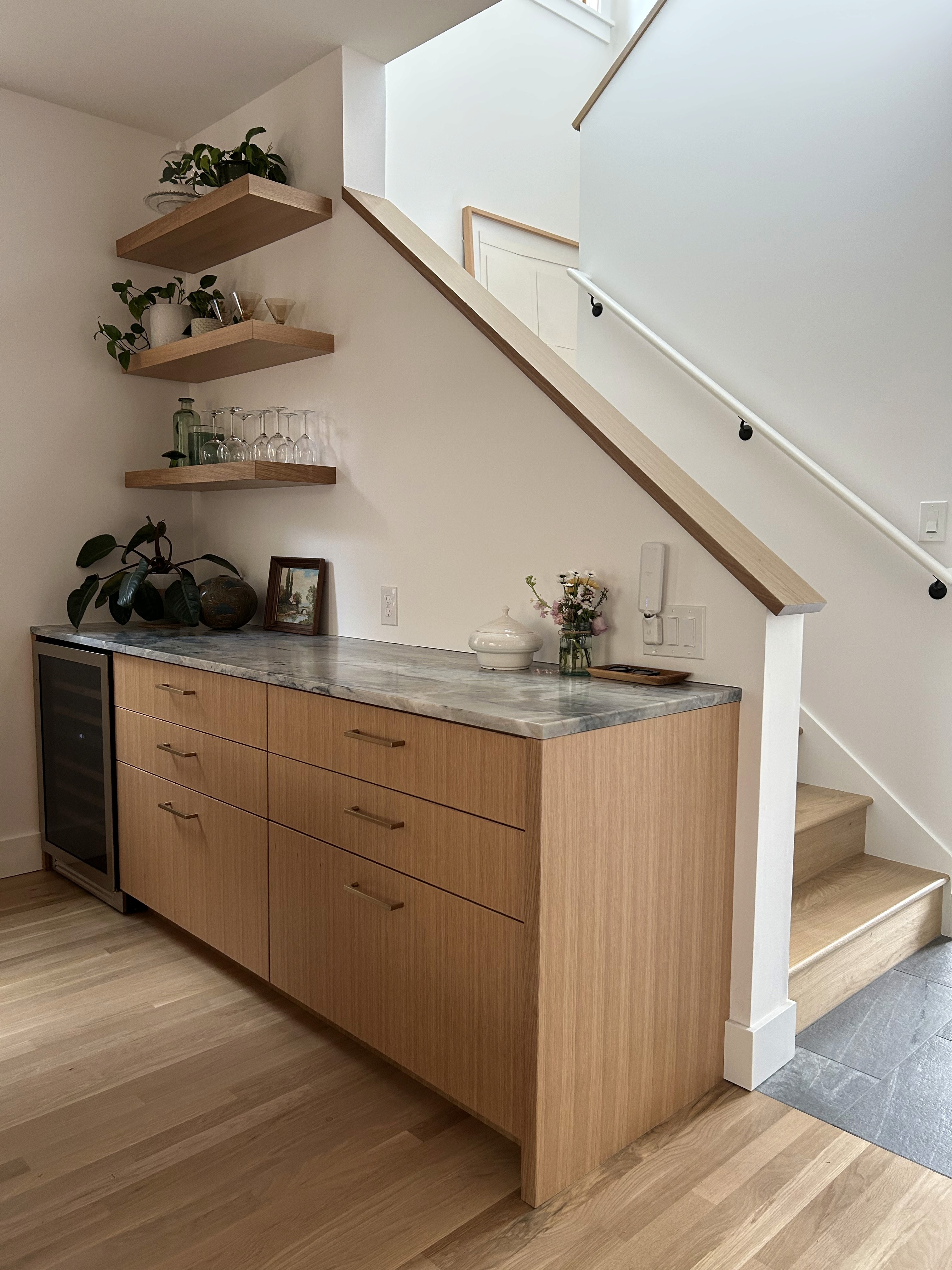 A white oak sideboard in a dining room sits below three wooden shelves with plants and next to an upper floor stairwell.