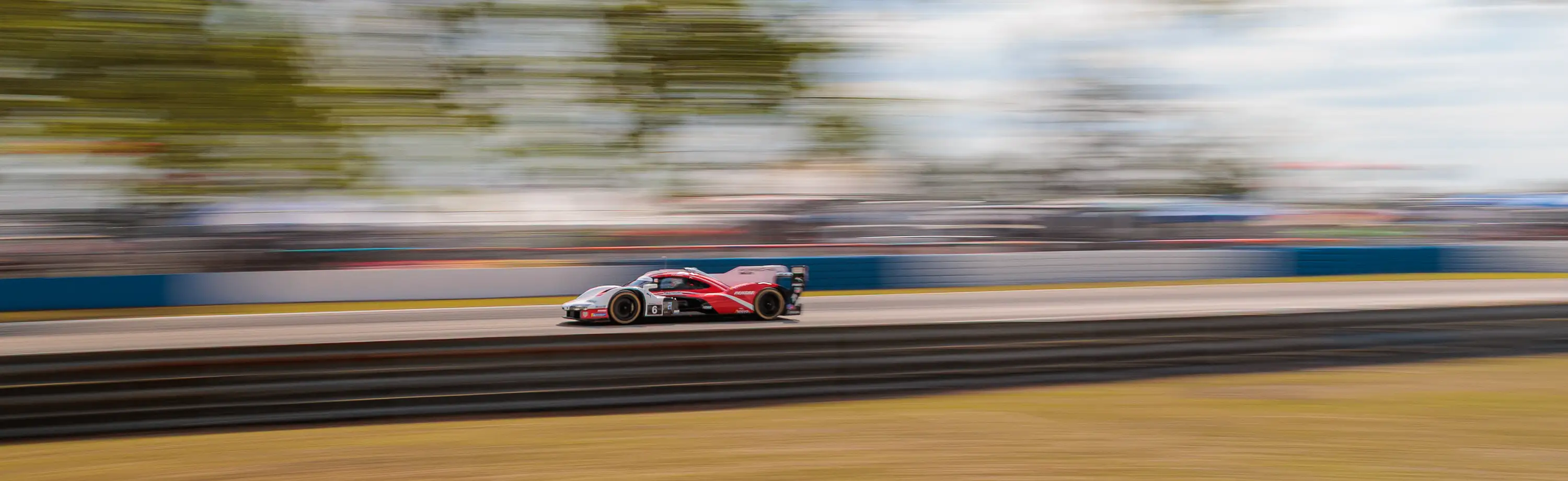 A red Porsche racing car speeds down a track, against a blue sky and trees, with a motion blurred background.