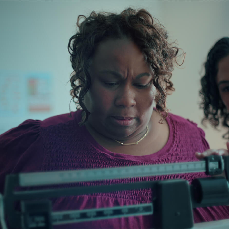 A woman in a purple top looks down at a mechanical weight scale, appearing focused, with another person partially visible beside her.