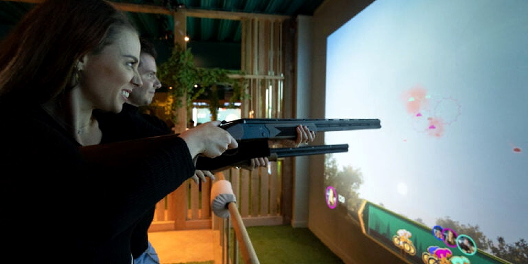 Two guests playing a digitally enhanced clay shooting game inside a hospitality venue with a large projection screen and social space around them.