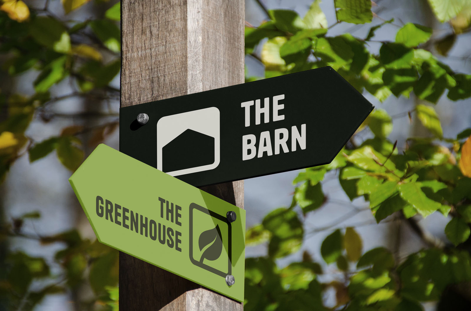 Een houten paal met twee pijlvormige wegwijzers in een groene, bosrijke omgeving. De bovenste pijl is zwart met een wit icoon en de tekst "THE BARN"; de onderste pijl is lichtgroen met een blad-icoon en de tekst "THE GREENHOUSE".
