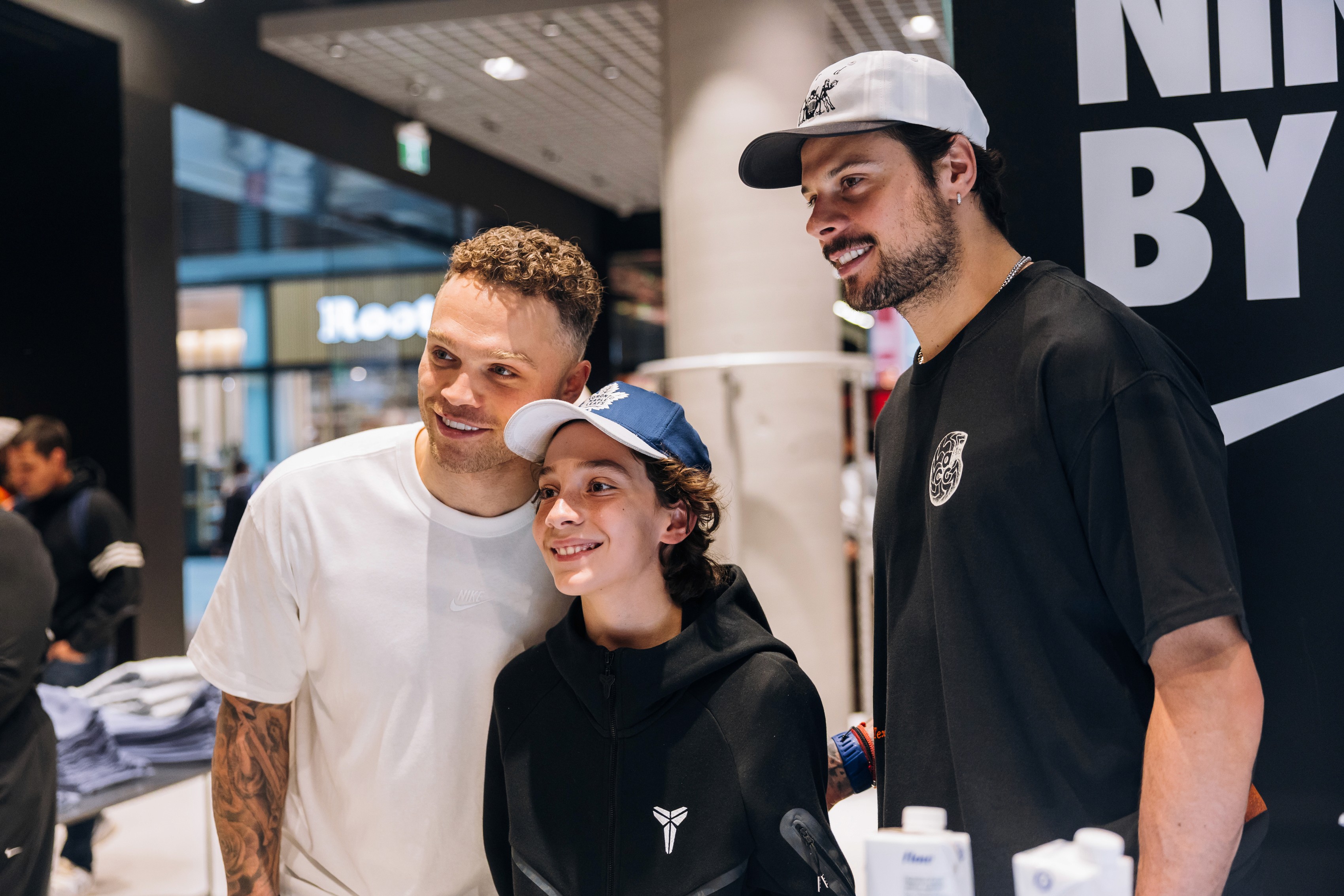 Three people smiling together in a store setting. Two athletes stand on either side of a child wearing a blue cap. They appear happy and relaxed.