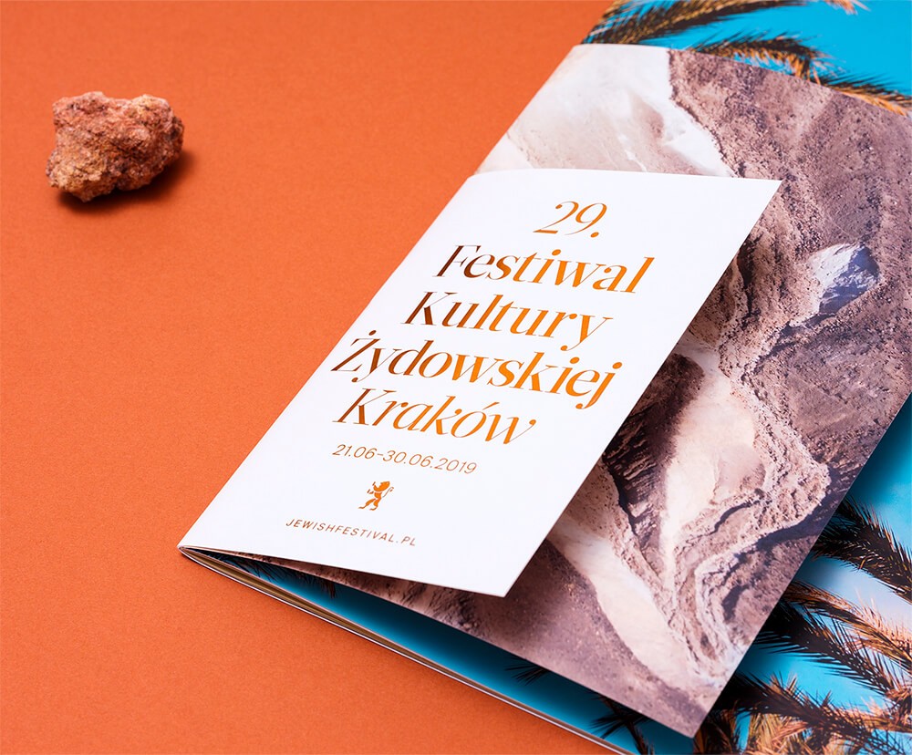 Biała okładka programu z pomarańczowym tytułem 29. Festiwalu leży na drukach ze zdjęciami skał i palm na pomarańczowym tle.