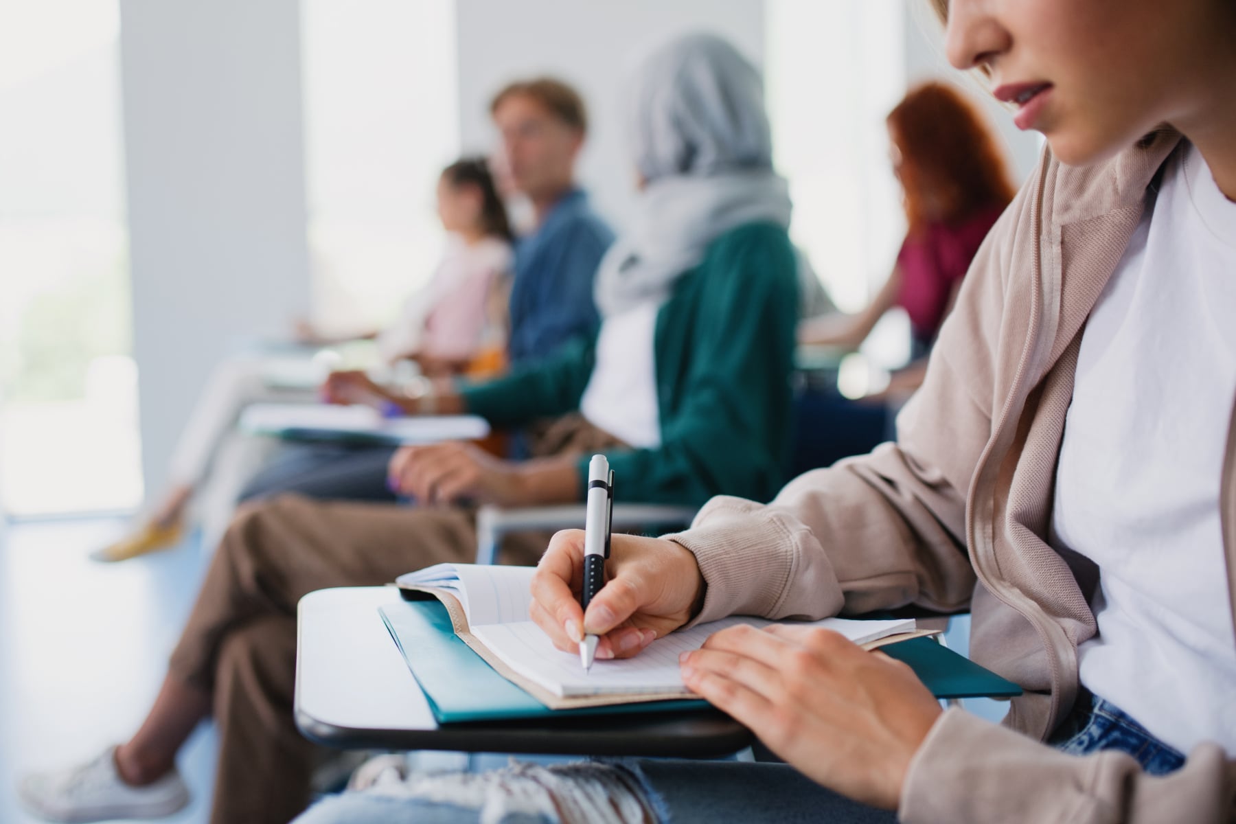 Students sit in a row in a bright classroom, with one student in the foreground writing notes on a clipboard.