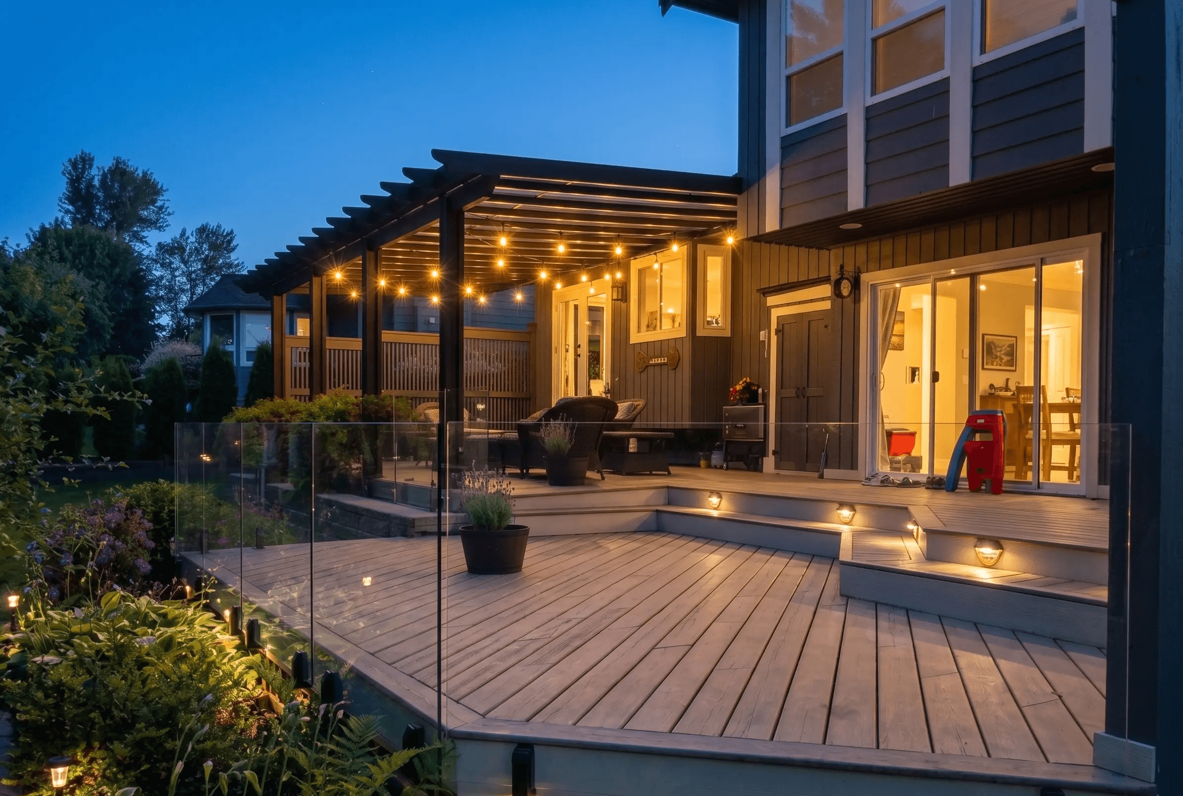 Frameless glass railing installed on a multi-level backyard deck in Duncan, BC, with pergola string lights and landscaped garden beds at dusk.