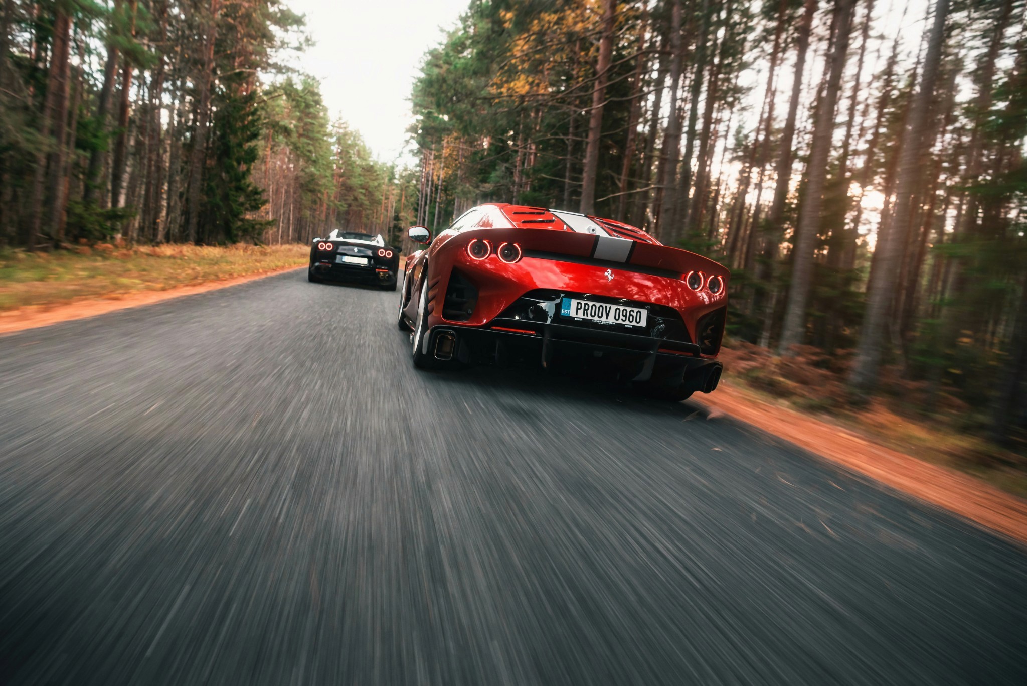 Two sports cars speed down a forested road, surrounded by tall trees, with the focus on a red car in the foreground showcasing sleek lines and modern design.