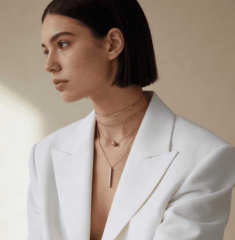 Minimalist fashion portrait of a young woman with sleek dark bob haircut, looking thoughtfully to the side; she wears layered gold necklaces (choker, chain, and a long bar pendant) over a structured white blazer with deep V-neckline, soft natural light illuminating her skin against a warm beige background.