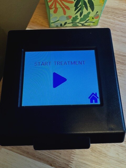 A small black device with a blue screen displaying a play button icon, placed on a surface.