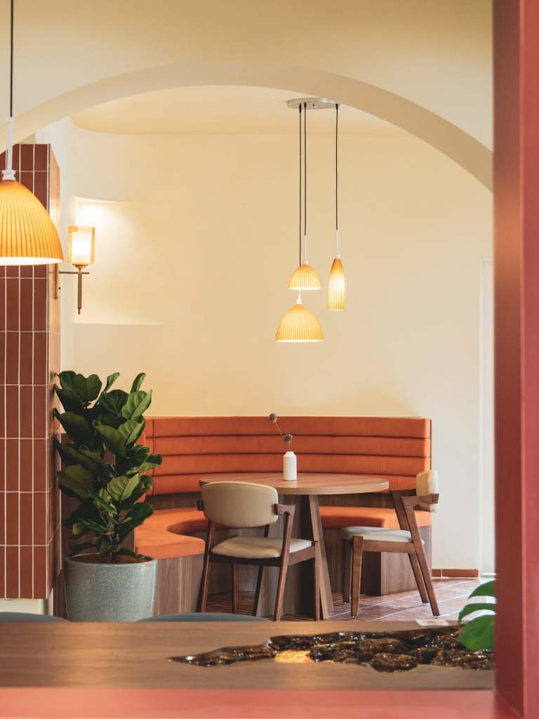 Cozy restaurant interior featuring an orange upholstered booth, a round wooden table with minimalist chairs, and pendant lighting against a light-colored wall.
