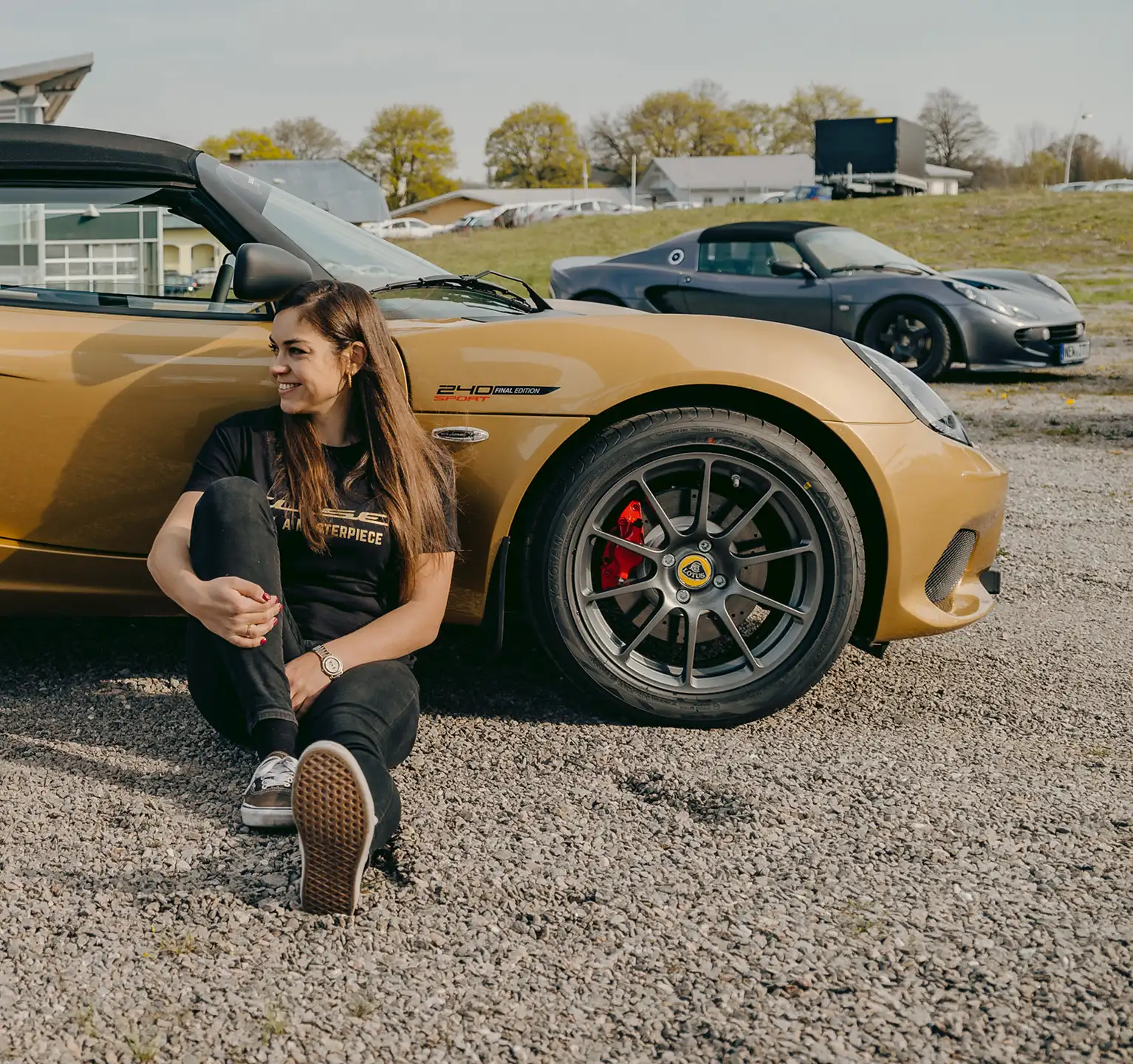 Elisa Artioli sitting on the ground, smiling next to a gold Lotus Elise 240 Final Edition parked on gravel, with another Lotus visible in the background.