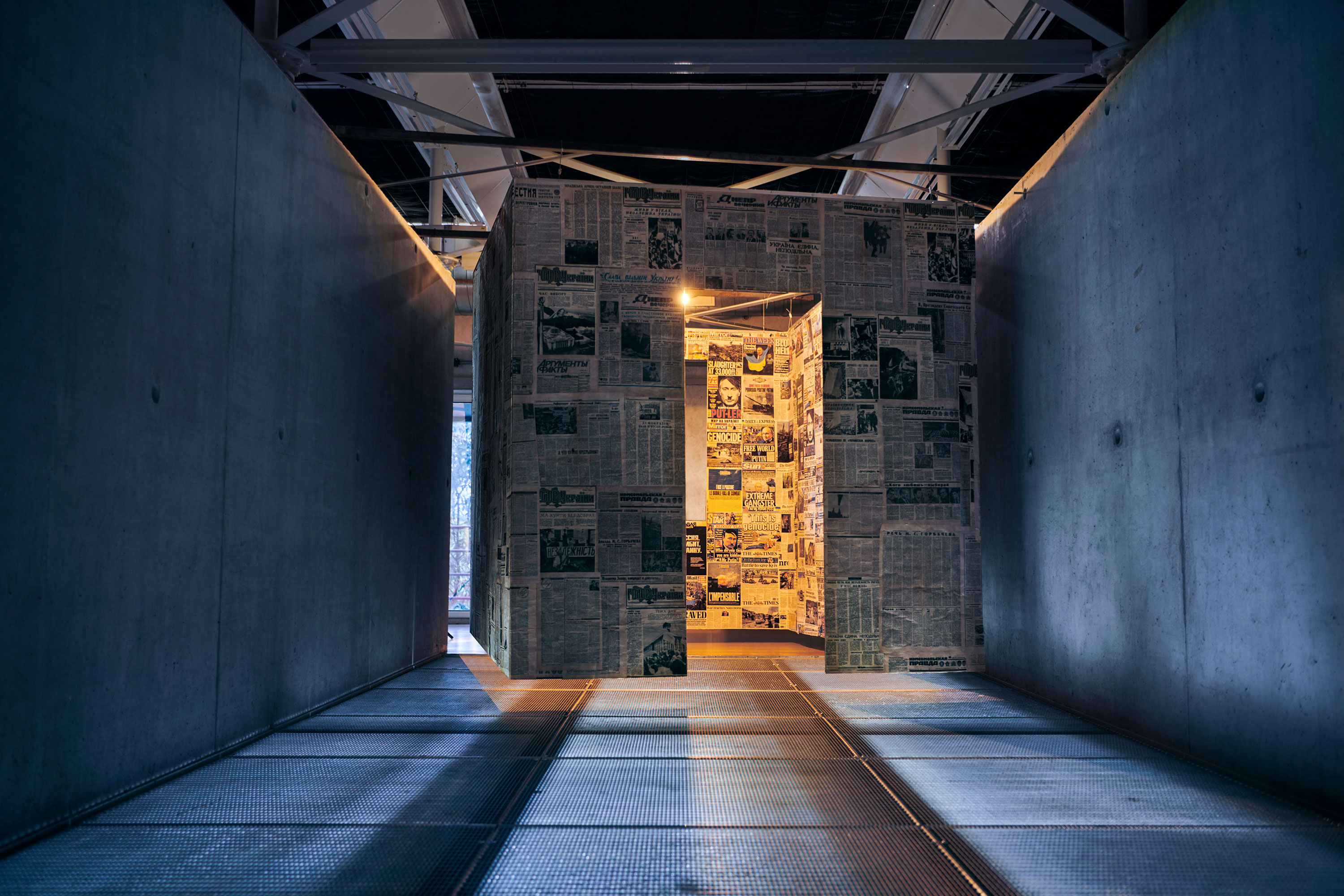 Front view of the newspaper-covered structure positioned between two concrete walls; a doorway cut into the surface reveals brightly lit walls inside covered with colorful newspaper front pages.