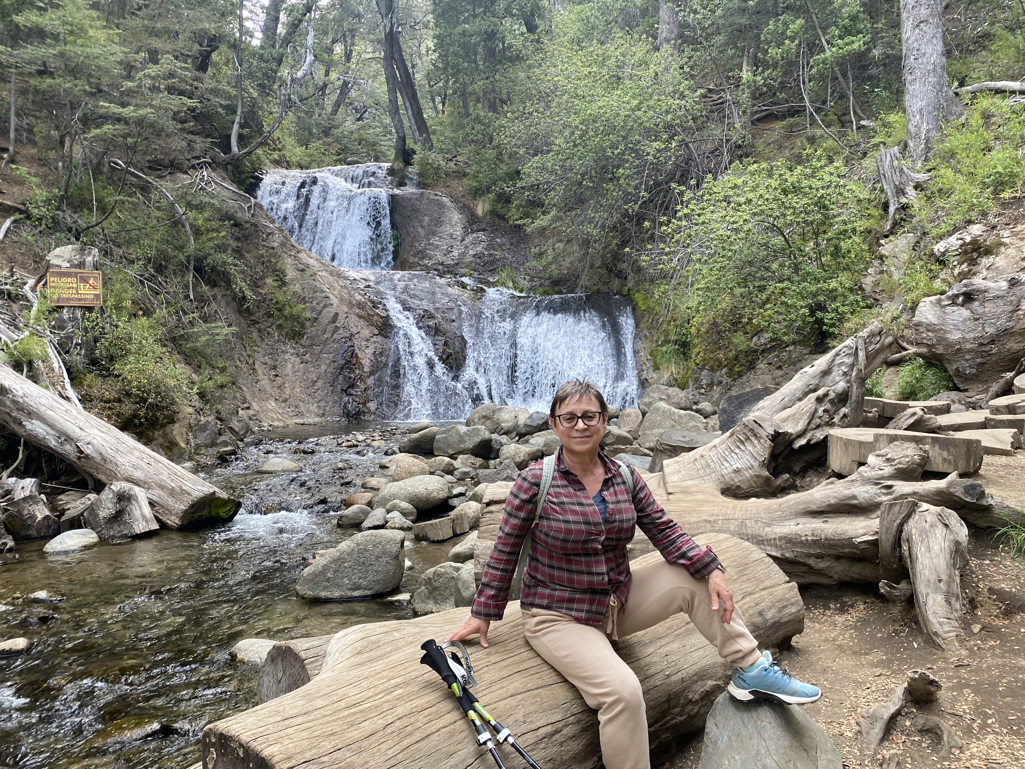 Mujer sentada sobre un tronco frente a una cascada doble en un bosque frondoso de Argentina.