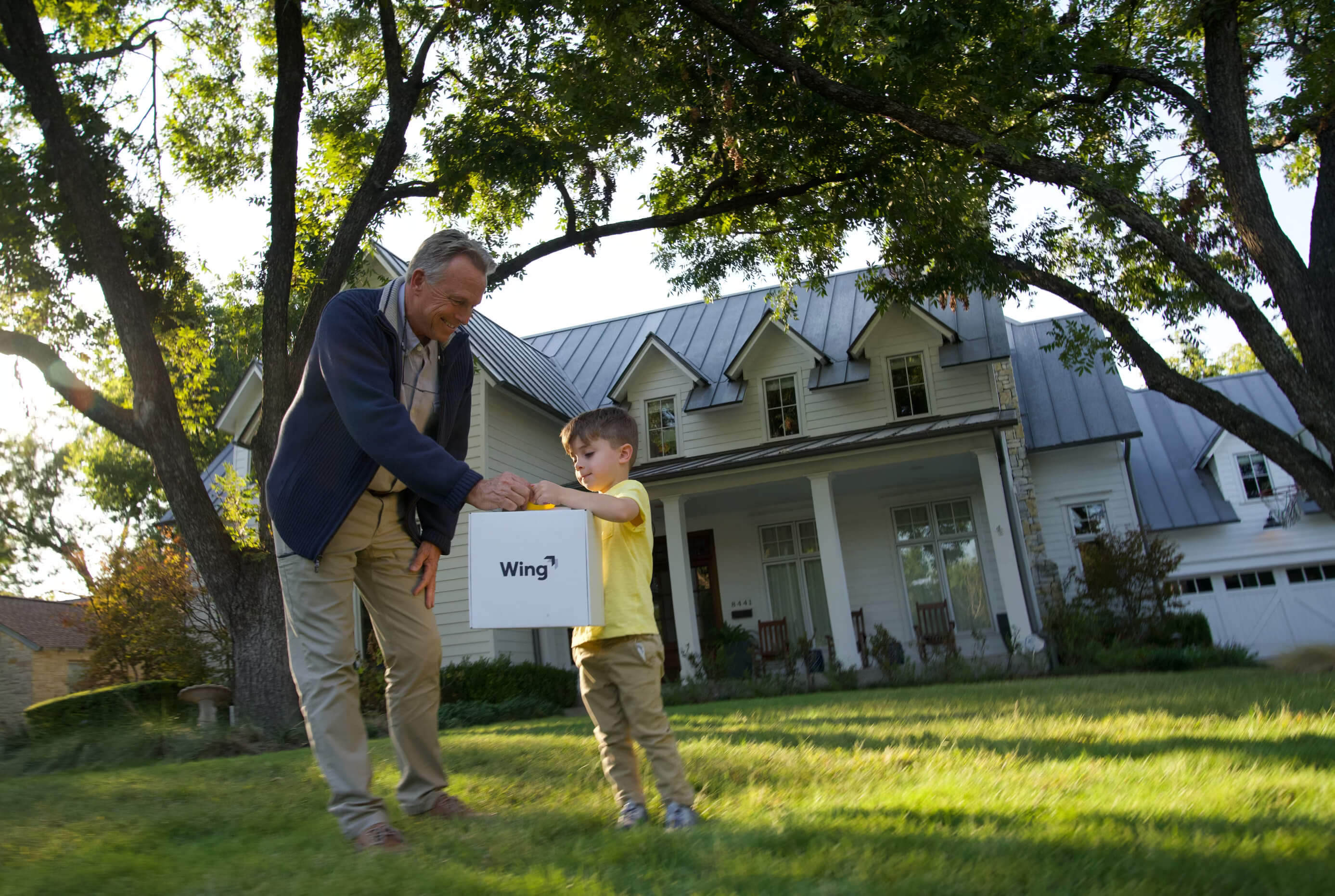 A grandfather and child stand on a lush green lawn in front of a large, charming house with tall trees, as they receive a drone delivery package marked "Wing".