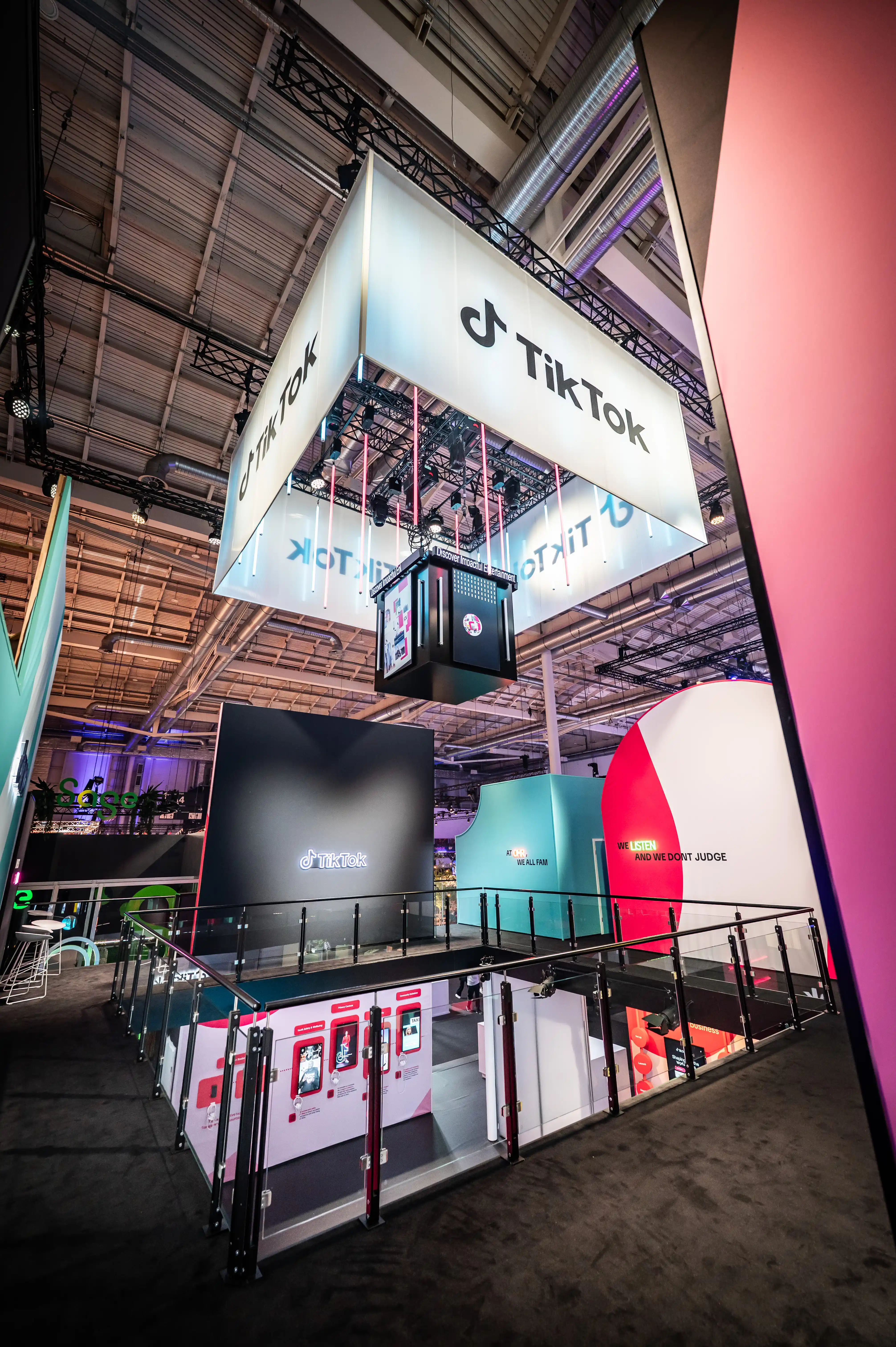 Tall two-storey TikTok booth with illuminated logo cube hanging above the upper deck in the exhibition hall.