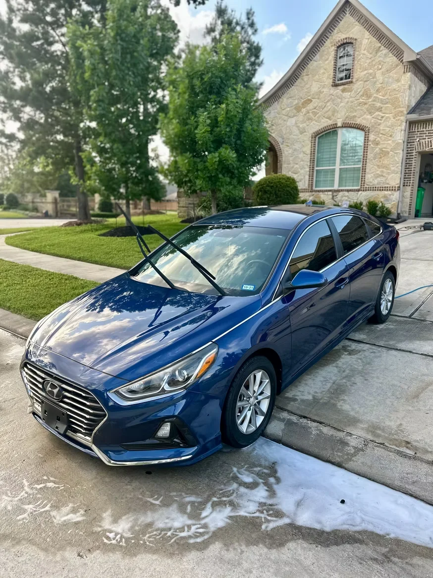 A dark blue Hyundai Sonata receiving a professional mobile car wash in a Houston driveway with soap suds visible on the ground.