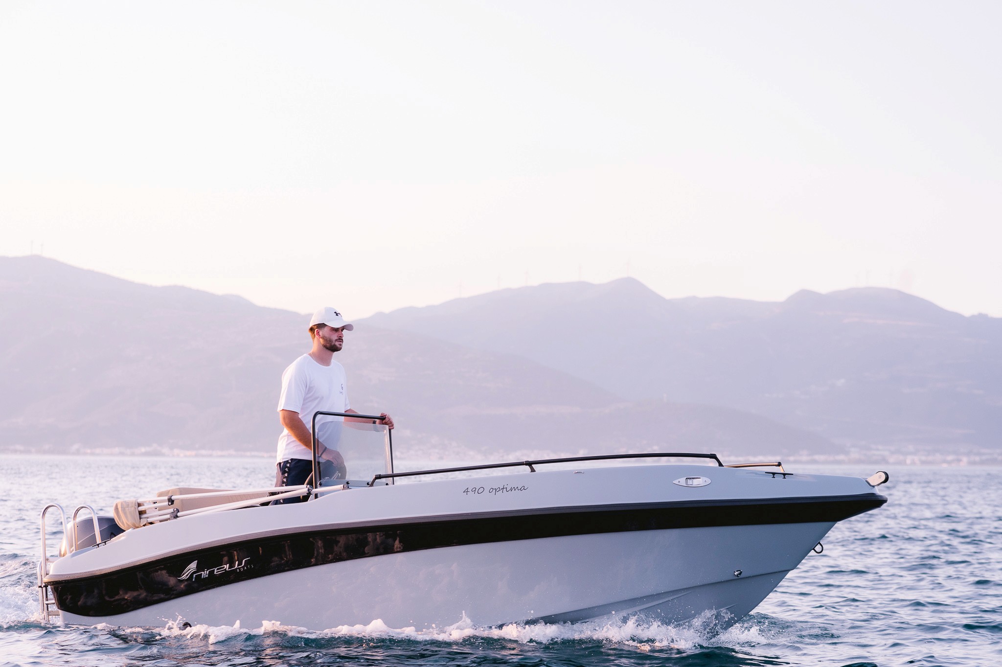 Captain piloting a sleek Nireus Optima Aura speedboat across blue waters with mountainous Greek islands in the background.