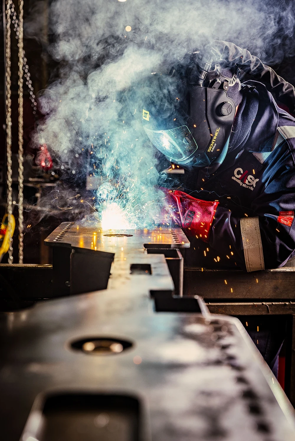 Welder carrying out MIG welding on a steel component in a fabrication workshop, with visible sparks and welding smoke.