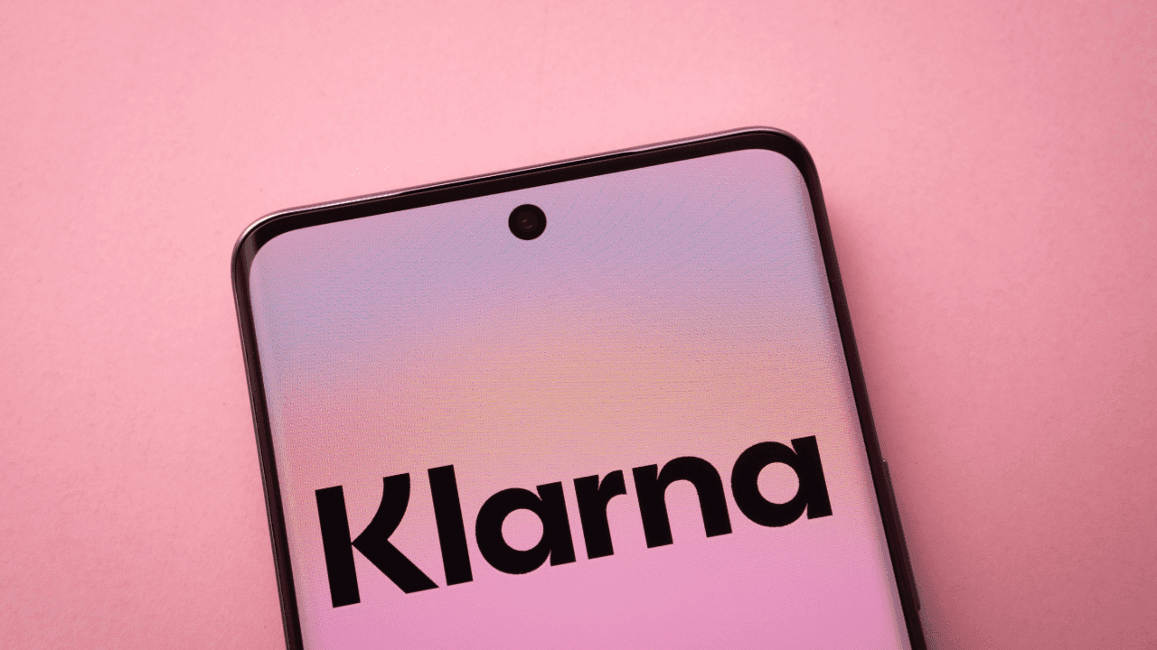 Smartphone in Nahaufnahme auf rosa Hintergrund, der Bildschirm zeigt „Klarna“. Sanfte Pastellfarben wirken ruhig und modern.