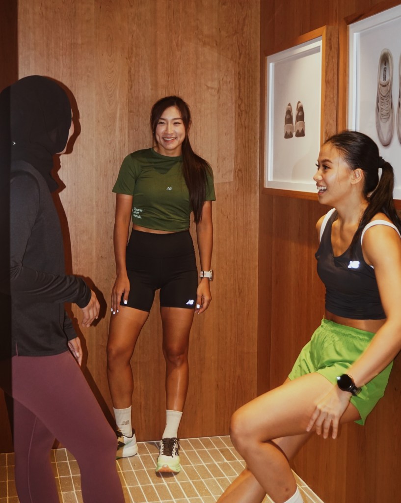 Three women engaged in conversation in a wooden room, wearing athletic clothing and smiling as they share a moment together.