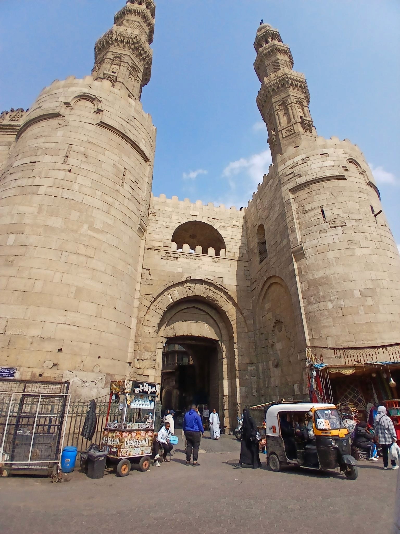 A gate from fatimi cairio, outside the gate are two large mnarets and some people and a rikshaw 