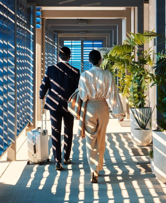 A couple walking through a sunlit corridor with blue louvers, carrying a suitcase, suggesting travel or vacation.