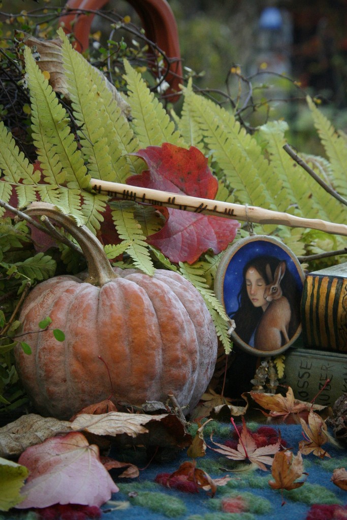 A rustic autumn arrangement featuring a pumpkin, vibrant fall leaves, fern fronds, and a small portrait of a woman with a rabbit, evoking a cozy, nature-inspired seasonal atmosphere.