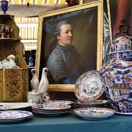 A table displays porcelain plates, vases, and figurines, with a framed portrait of a man in the background.