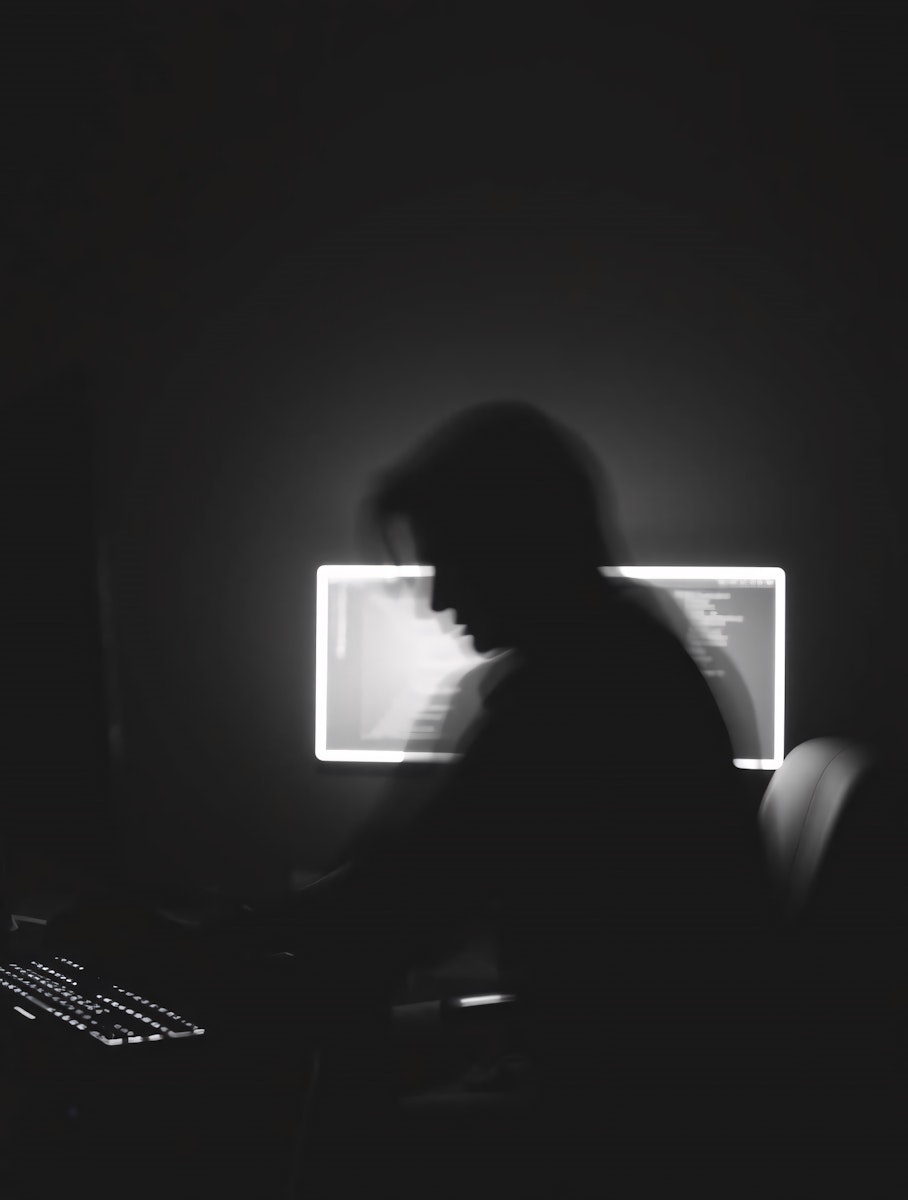 Blurry silhouette of a person working at a computer desk in a dark room.