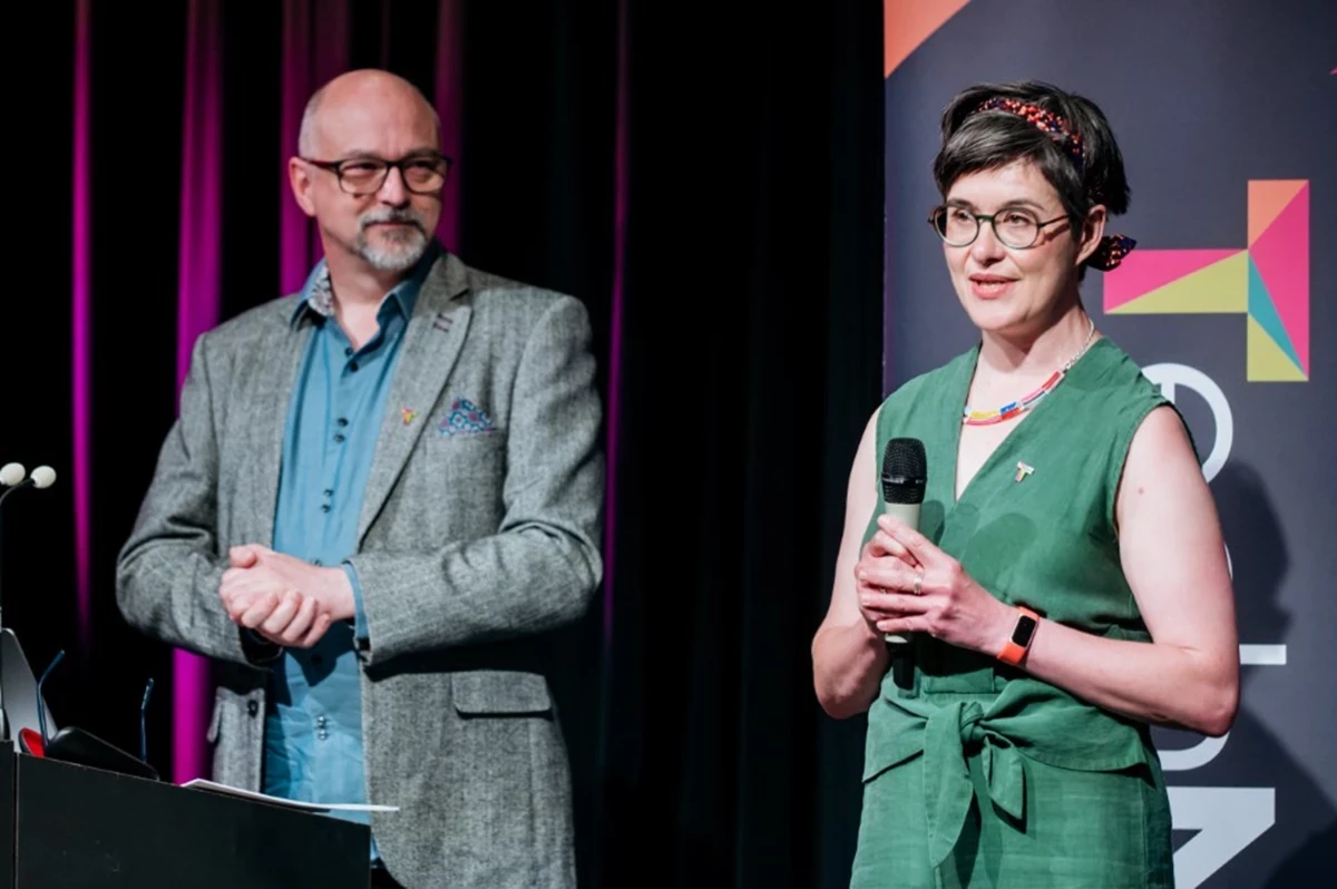 Two people standing on stage in front of a dark backdrop with colorful accents; one is holding a microphone, and the other stands near a laptop on a podium.