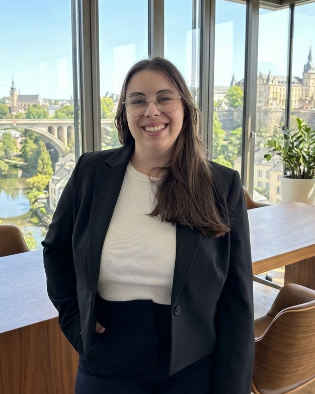A young man in a formal outfit with a dark overcoat and rust-colored vest, gazing directly at the camera against a neutral background.