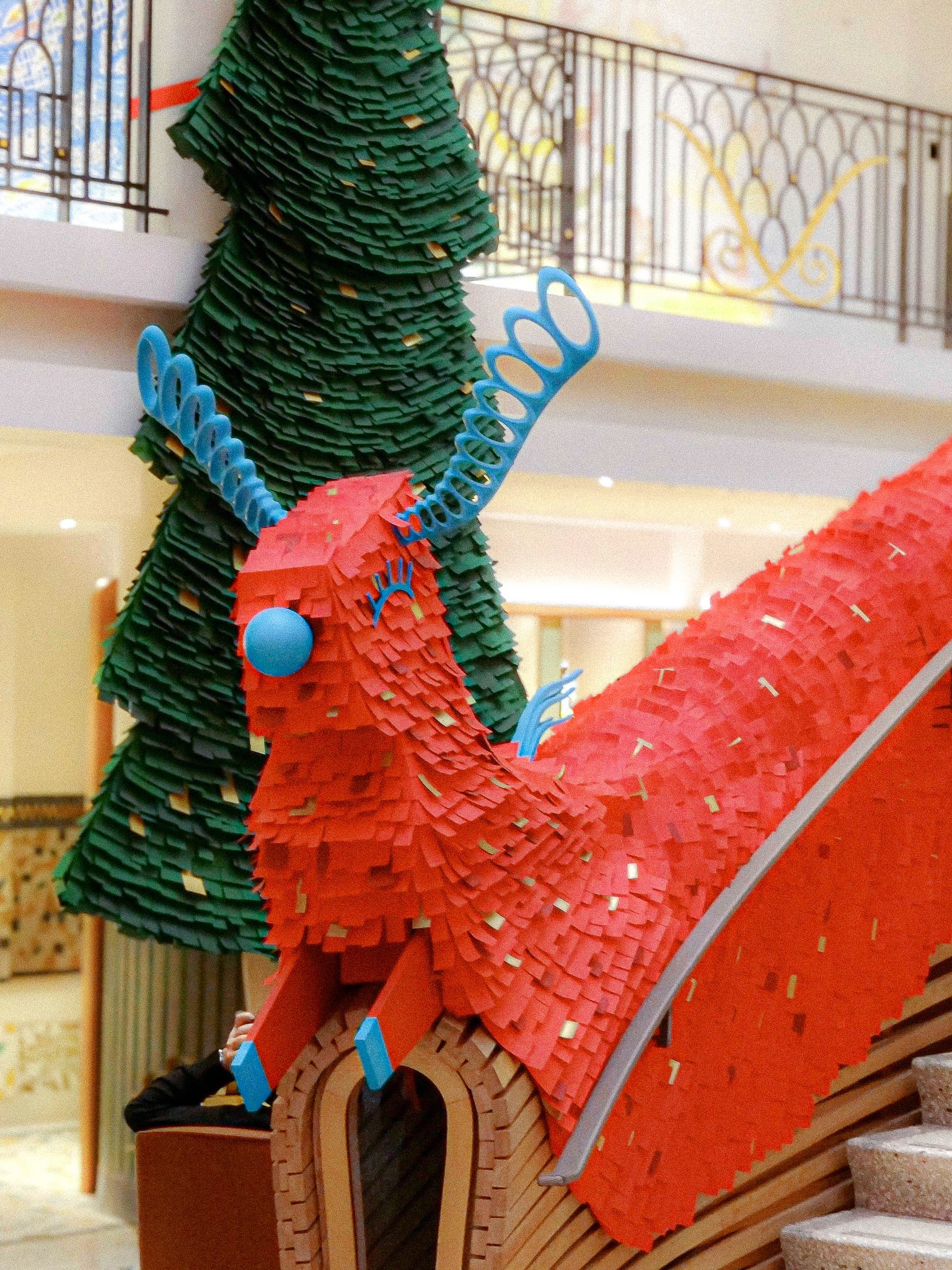 In-store holiday installation for Hermès featuring a large red fringed reindeer character sliding down a staircase banister next to a stylized green paper Christmas tree, designed by WM Paris.