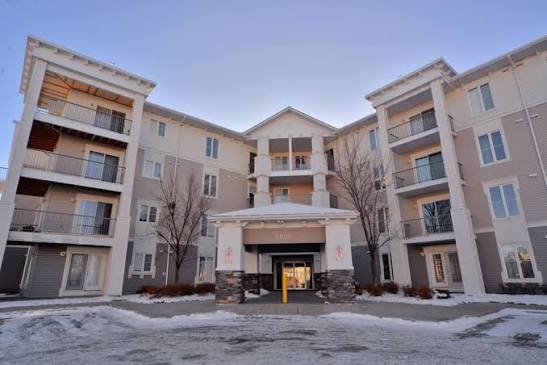 Four-story residential condominium building featuring light grey siding, a covered central entrance with stone pillars, private balconies, and a snow-dusted driveway in Taradale, NE Calgary