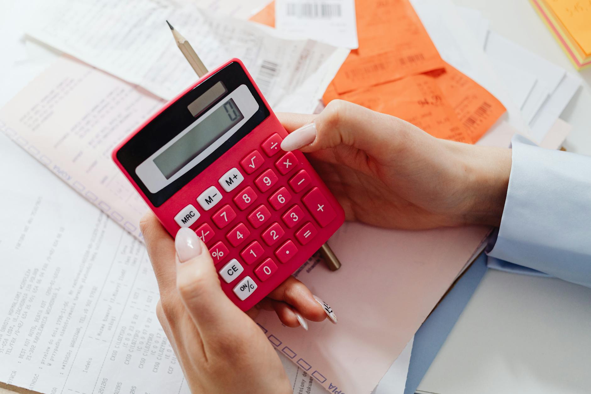 Hands using a red calculator surrounded by financial receipts and paperwork on a desk