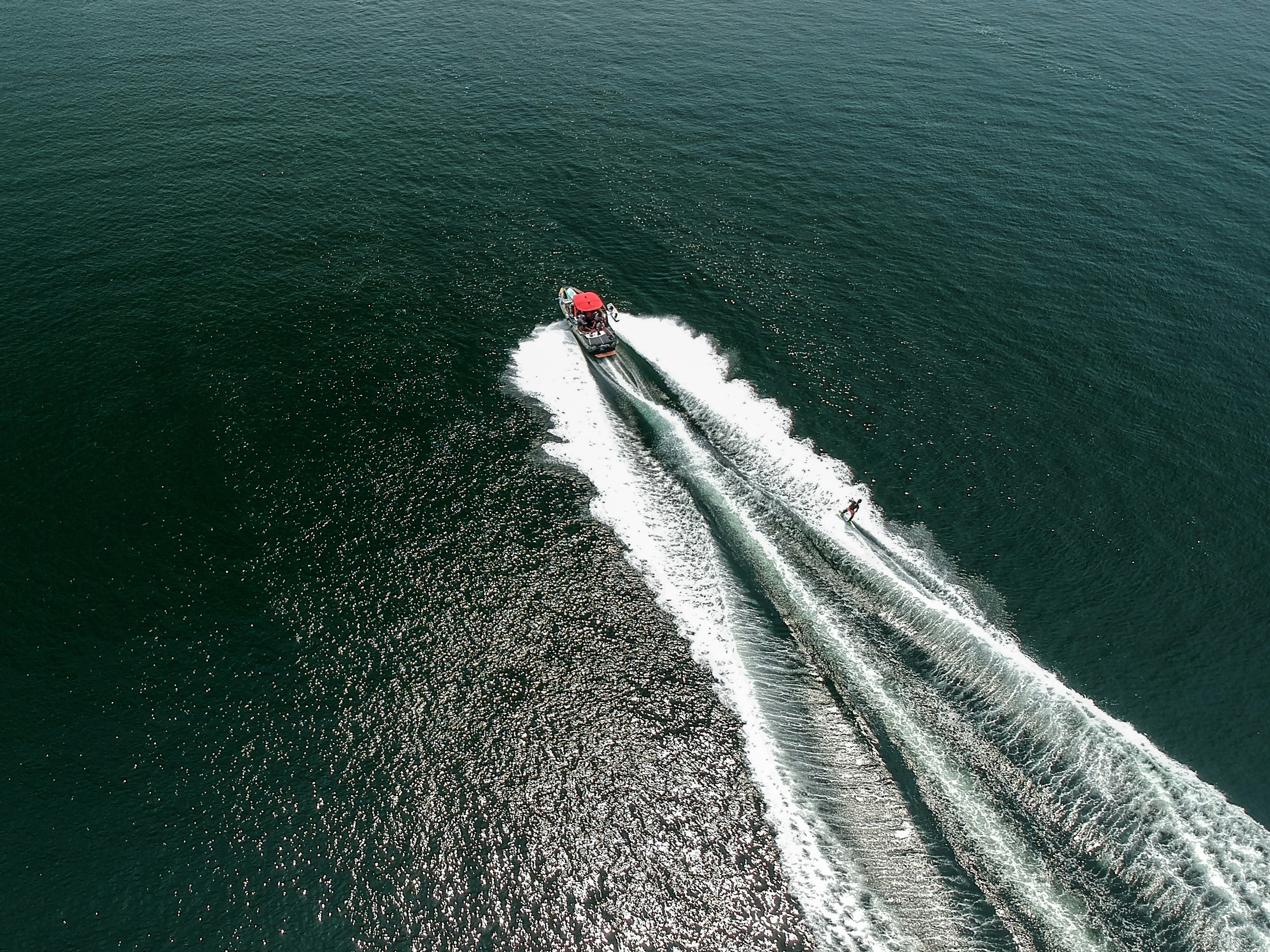 Aerial view of a speedboat with a bright red canopy creating white wakes as a person water skis behind it on a vast, deep blue ocean expanse.