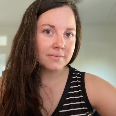 A woman with long, brown hair and a striped black and white tank top looks directly at the camera, set against a softly blurred indoor background, conveying a calm and approachable demeanor.