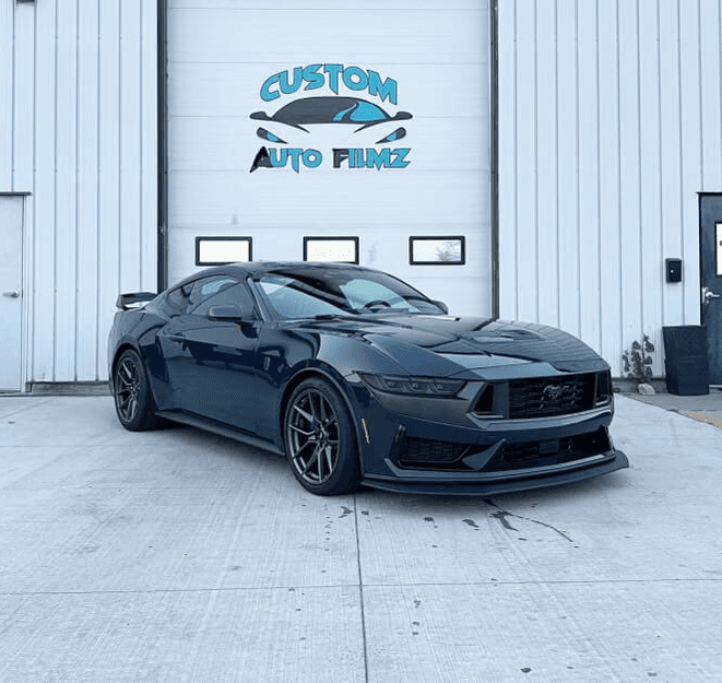 A dark blue Ford Mustang Dark Horse is parked on a concrete driveway in front of a white industrial building.